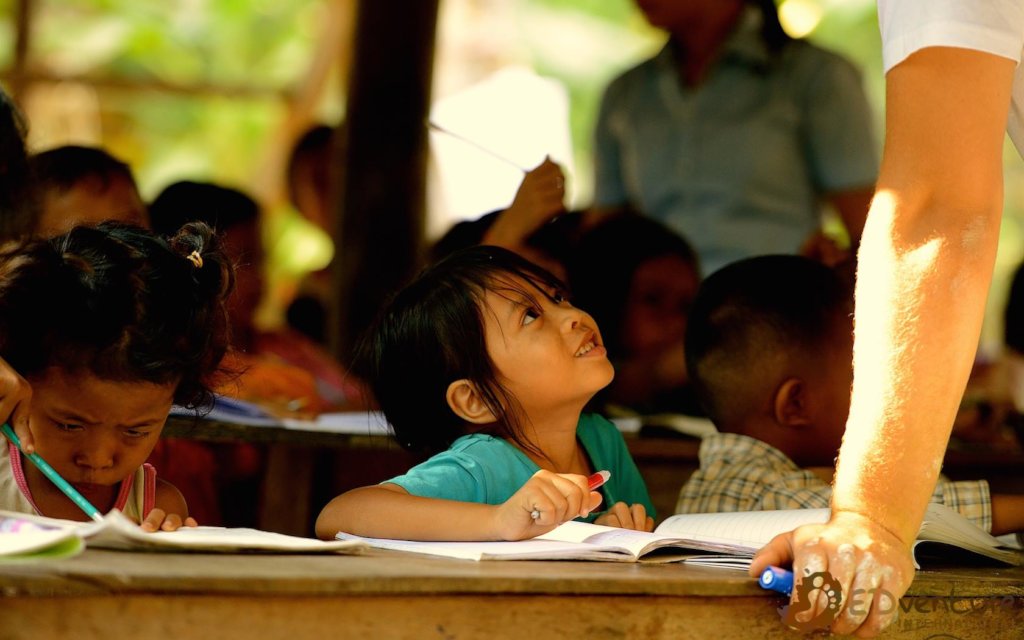 EDGE Worldwide Cambodia educational charity — students and educators in the classroom A student looking up at an adult beside her desk in a Cambodian classroom